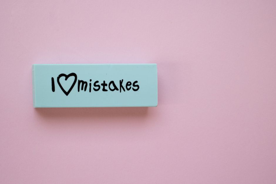 A light blue rectangular sign with the phrase 'I ♥ mistakes' written in black ink, placed on a soft pink background. The sign features a heart symbol replacing the word 'love,' emphasizing a positiv